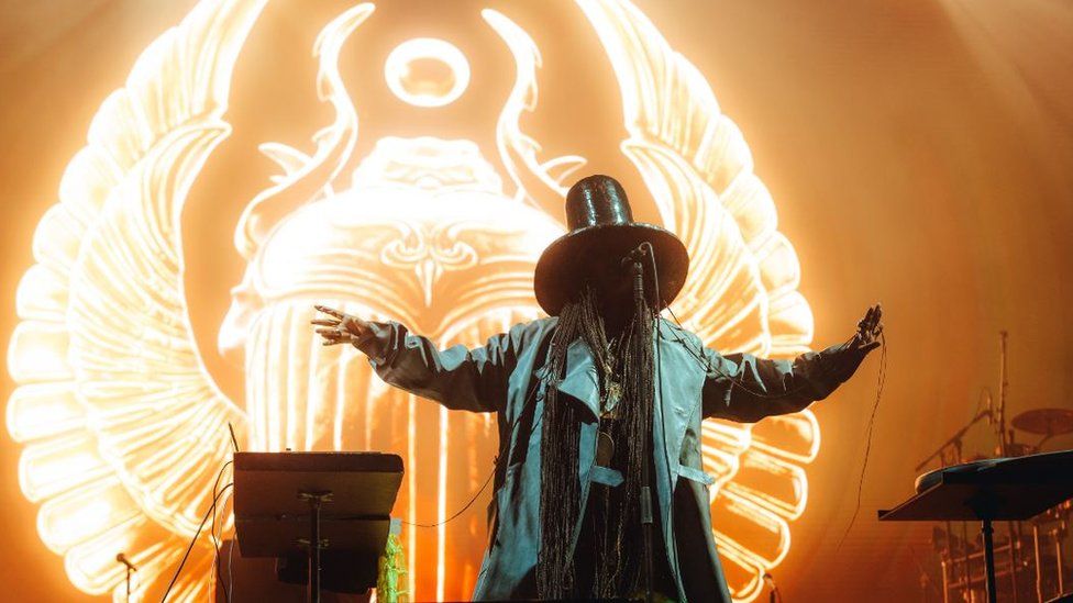 An artist in a large hat and cape on a stage