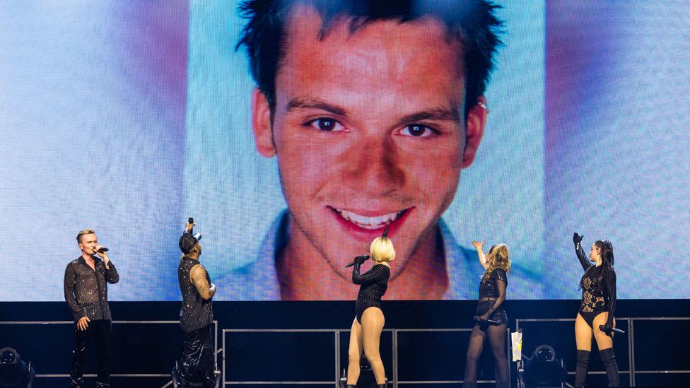 S Club on stage at Manchester AO Arena in front of a large photo of Paul Cattermole on the big screen