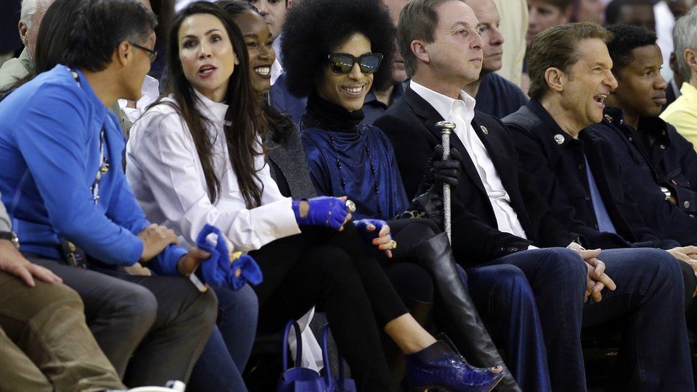 Singer Prince, in sunglasses, smiles as he watches an NBA basketball game between the Golden State Warriors and the Oklahoma City Thunder on Thursday, March 3, 2016, in Oakland, California