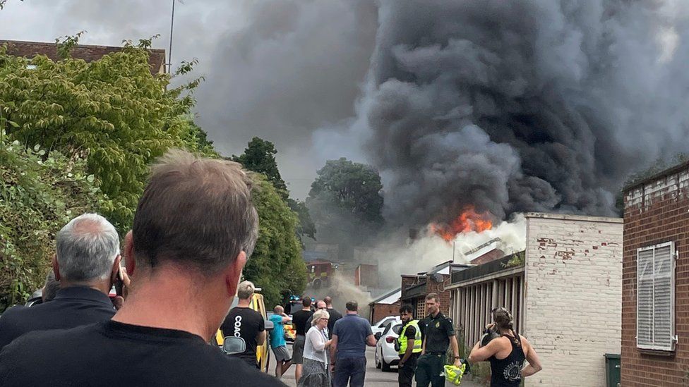 Plume of smoke above car workshop fire, with people looking on