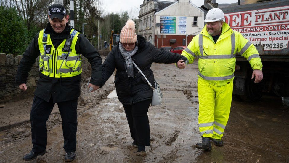 A woman helped through mud in Mountain Ash, Rhondda Cynon Taf, by a police officer and a council worker