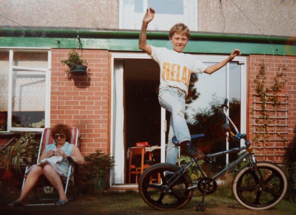 son shows off a balancing trick on the bike while woman knits in background