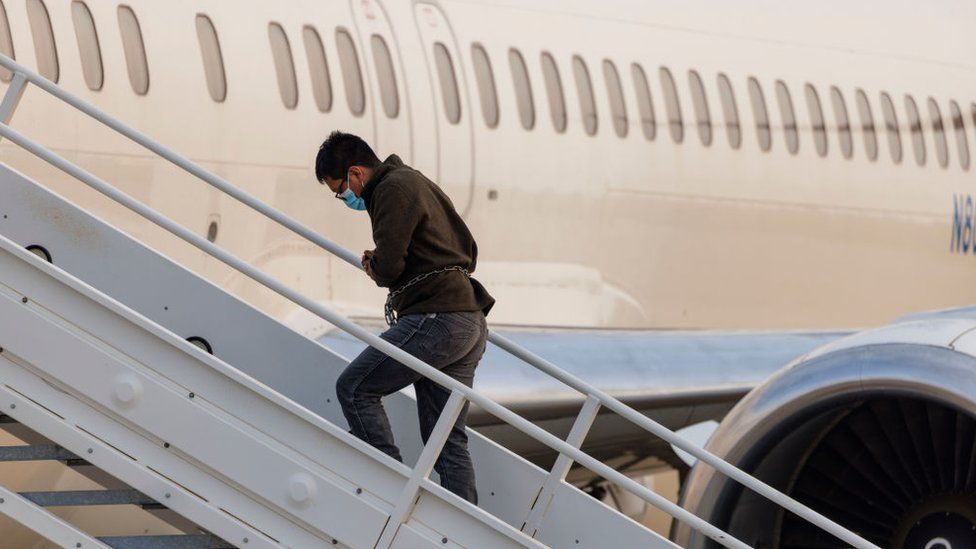 A shackled migrant boards an airplane