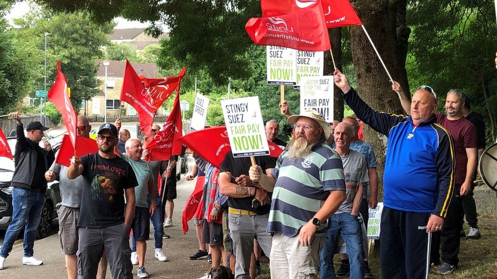 Seveal men holding red Unite flags and placards saying 'Stingy Suez Fair Pay Now'