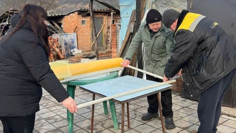 Two men and a woman, standing around a table, build an Insulate Ukraine window