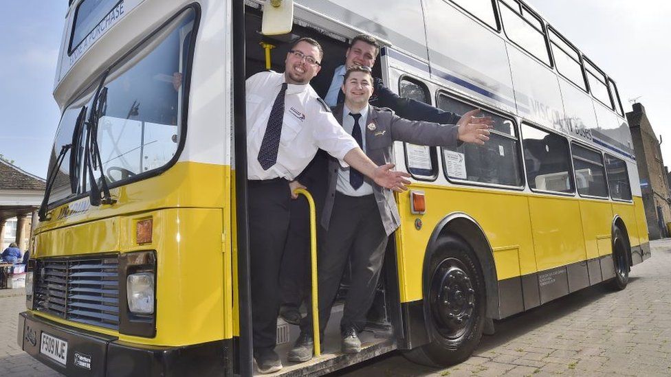 Nathan Merryweather and two colleagues wave while standing at the entrance to a bus