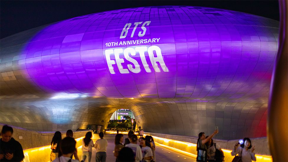 People gather to celebrate the tenth anniversary of BTS at the Dongdaemun Plaza in Seoul, South Korea. Fans are taking pictures with a landmark building covered in purple, with a sign that reads "BTS 10th Anniversary".