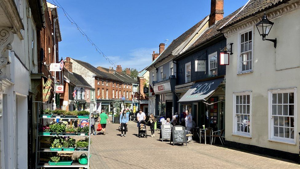 View down the main street in the centre of Halesworth. It is a sunny day and in the middle is a family walking with a pushchair towards the camera. On the left is a stand with various fruit and vegetables for sale. The buildings are a mix of Georgian buildings, some of which are red brick fronted and others have been smoothly rendered
