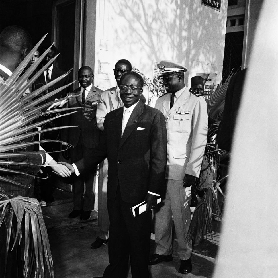 Security personnel flank Léopold Sédar Senghor, then-president of Senegal, who shakes hands with an unseen man.
