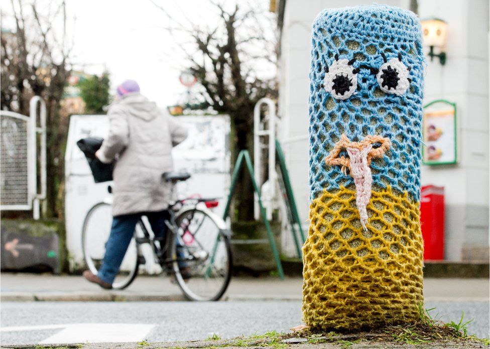 A concrete bollard in Hannover, Germany, is seen covered in bright knitted fabric with two eyes a a mouth - an example of yarn bombing