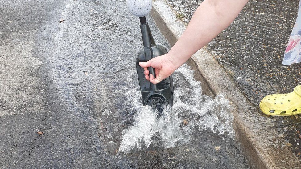 Neighbour puts watering can into stream of water on road