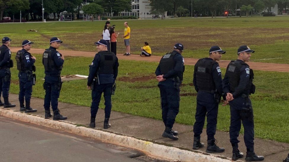 Brazilian riot police stand guard as two Bolsonaro supporters in yellow shirts are interviewed by journalists in Brasília. Photo: 11 January 2023