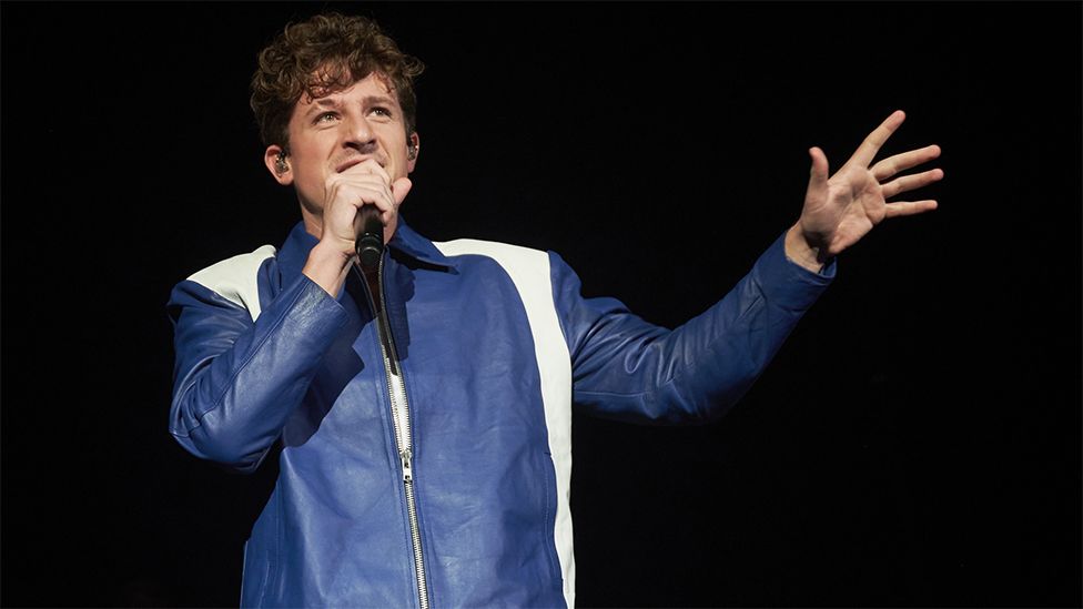 Charlie Puth holding a microphone to his mouth, with his left arm outstretched. He is wearing a blue jacket with white stripes running down the side. The background is plain black.