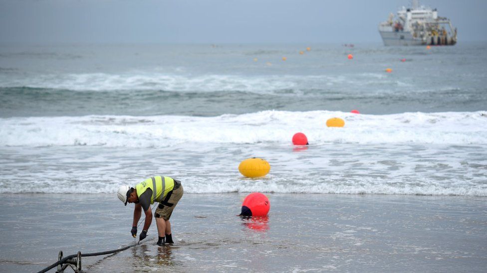 A worker on the edge of the sea with a ship offshore works on the mooring of a fibre optic cable seen rising out of the sea, that runs between the US and Spain in photo taken in 2017