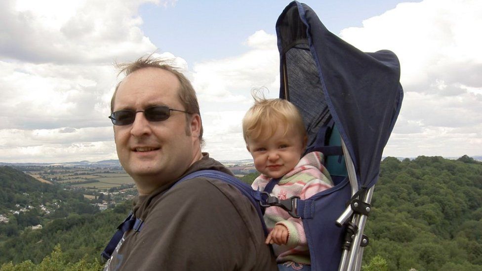 Abbie and her dad Andrew picture when she was a toddler, it looks like they are out in the countryside perhaps at the top of a hill. Andrew is standing smiling for the camera wearing sunglasses and a brown T-shirt. He is carrying Abbie on his back in a rucksack style baby carrier. Abbie is facing her dad's back and is looking at the camera. She has blonde hair and is wearing a pink, green, red and white stripey fleece top.