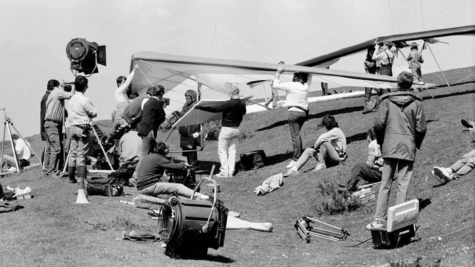 Black and white image of a television crew on a hillside surrounding David Jason in a hang-glider