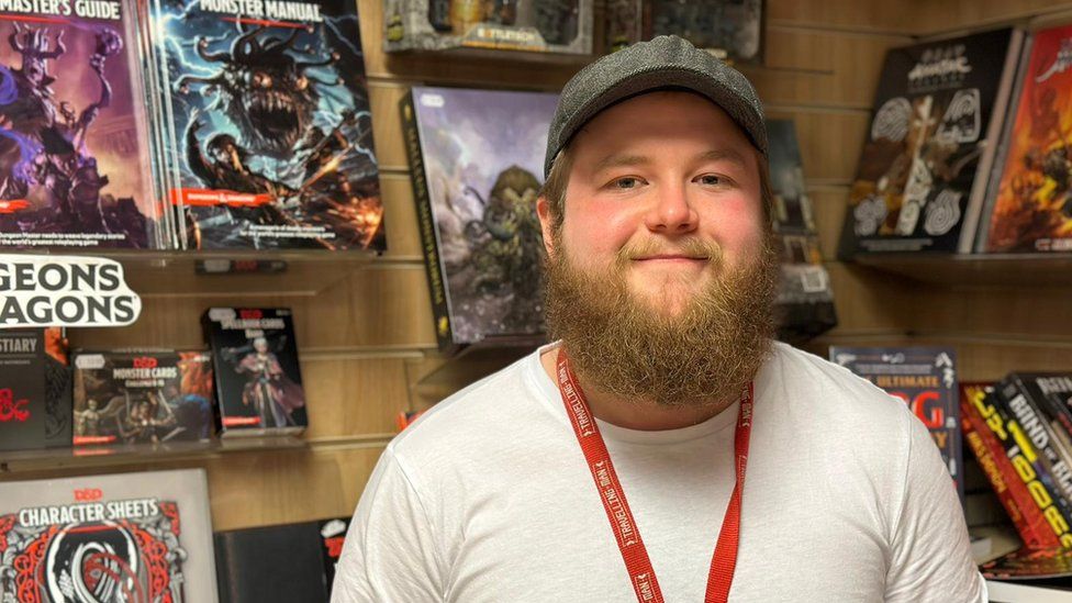 A man wearing a white t-shirt and a flat cap and a ginger beard smiles at the camera in front of shelves with Dungeons and Dragons books on them