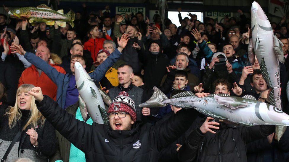 Grimsby Town fans cheering with several people holding inflatable Harry Haddocks
