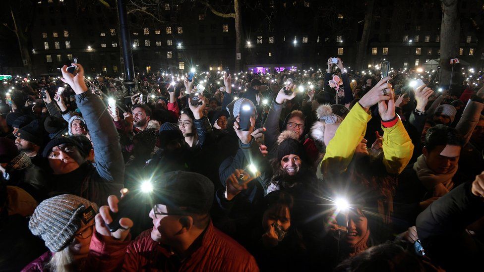 Crowds of happy people in winter clothing hold up their phone flashlights to get pictures of the display