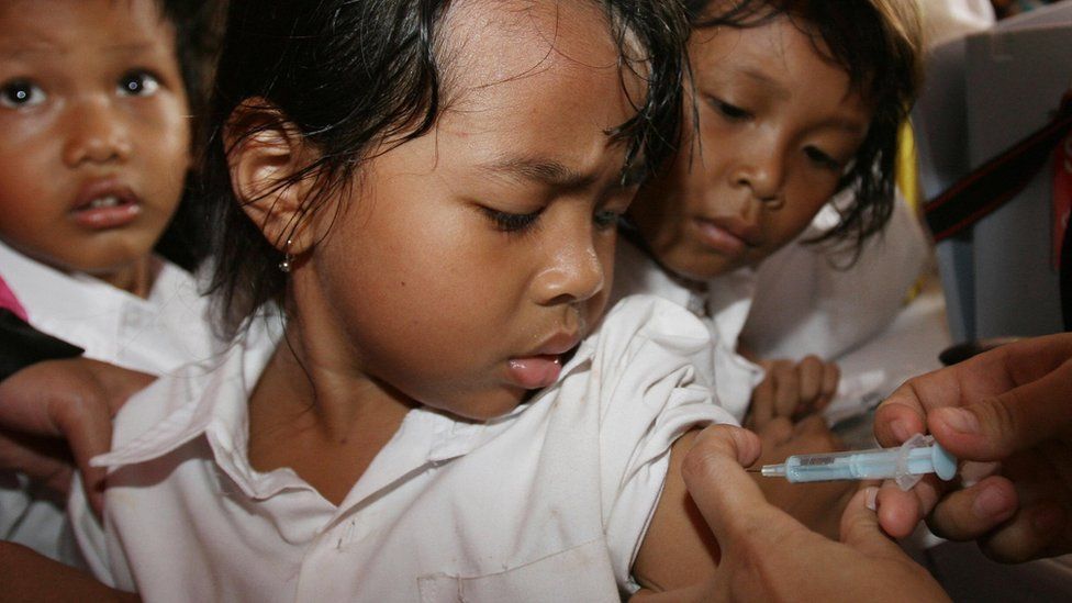 Girl watches as an adult hand administers a vaccine by needle into her upper arm. Child next to her also watches