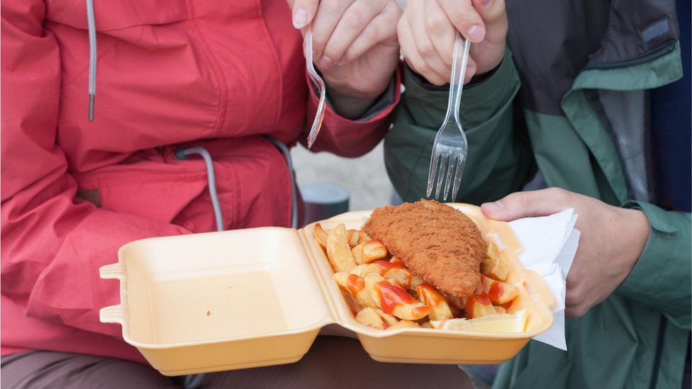 Couple eating fish and chips using a plastic fork from a single-use tray.