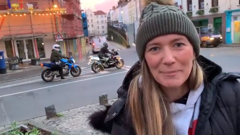 Lady standing in Guernsey town with two motorbikes in traffic in the background