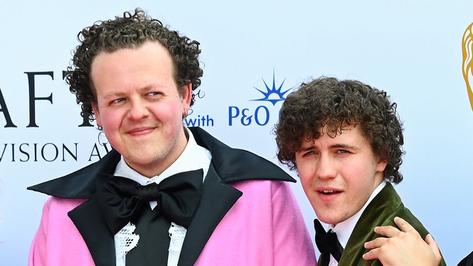 Jack Rooke, wearing a bright pink jacket with an oversized black collar, and Dylan Llewellyn, wearing a dark green dinner jacket over a white shirt with black bow tie, pose in front of a BAFTA hoarding on the red carpet