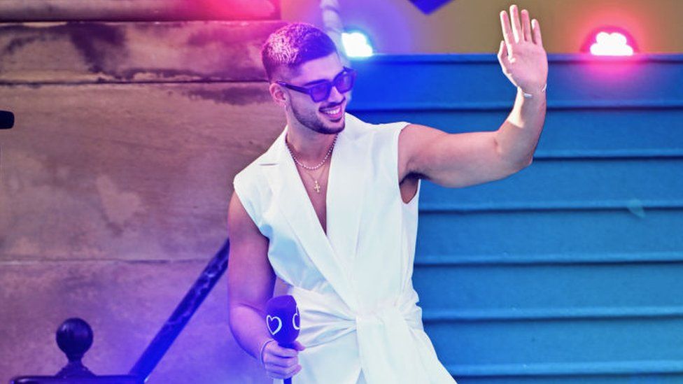 Andrew Lambrou wearing a white vest waving to the crowds and holding a microphone at the Eurovision turquoise carpet event in Liverpool