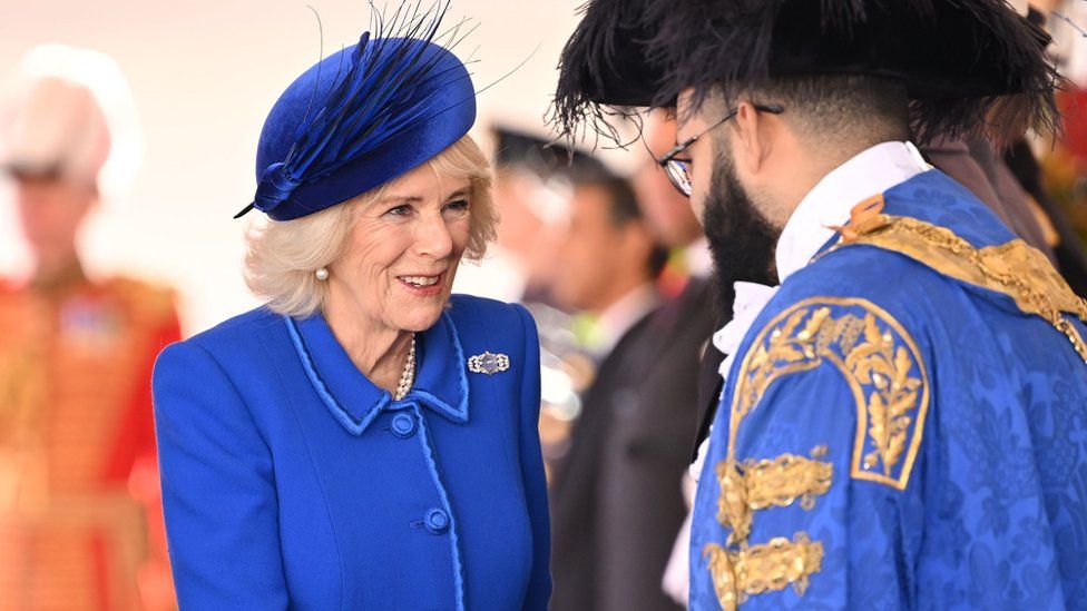 Queen Consort Camilla in a royal blue suit and hat, meeting with a man dressed in gold and blue robes.