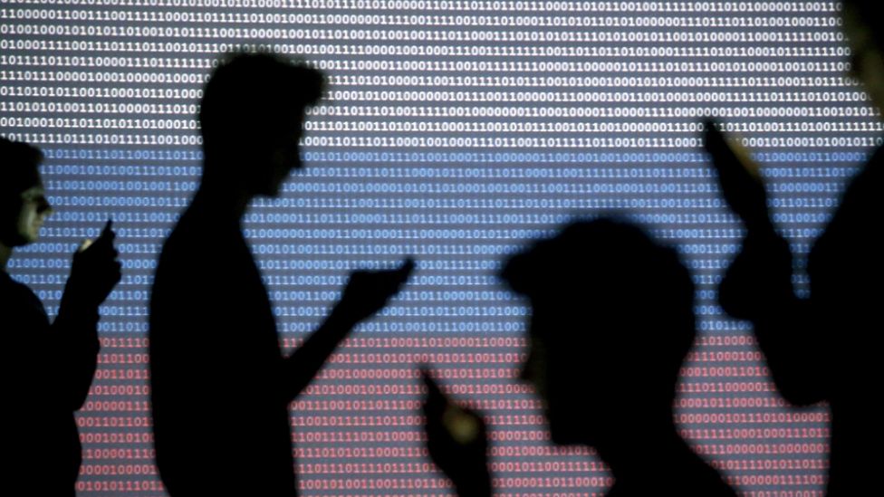 People are silhouetted (in silhouette) as they pose with mobile devices (mobiles, checking message, texting, sending texts, social media) in front of a screen projected with binary code and a Russian national flag