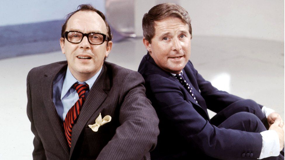 Eric Morecambe and Ernie Wise pose while sitting down