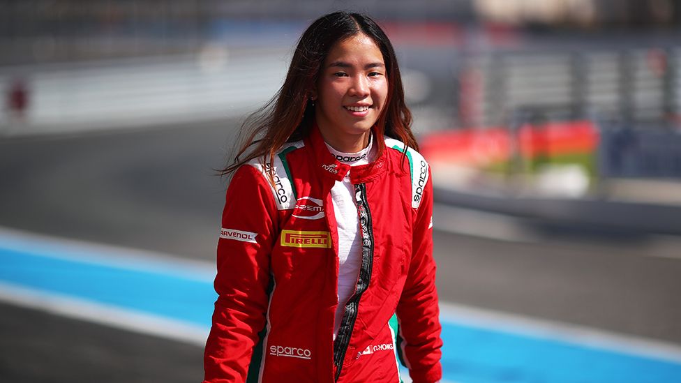 Chloe smiling, wearing red race overalls on a race track, with a white top. The overalls have advertising writing on the shoulders and across the front. The background is slightly blurred, with a streak of blue visible.