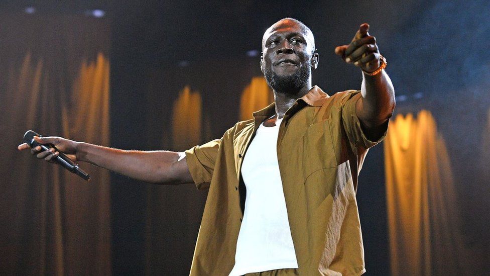 Stormzy performs at All Points East Festival 2023. Stormzy is a young black man with a shaved head and short beard. He wears an unbuttoned olive green shirt over a white T-shirt. With his left hand he points to the crowd while holding a mic outstretched in his right hand.