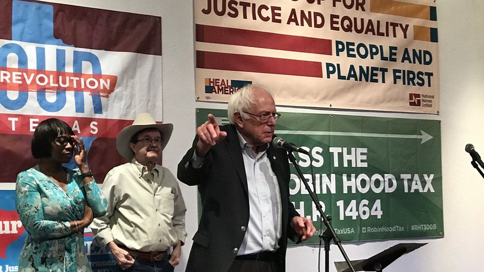 Bernie Sanders stands in front of a microphone, pointing as he speaks.