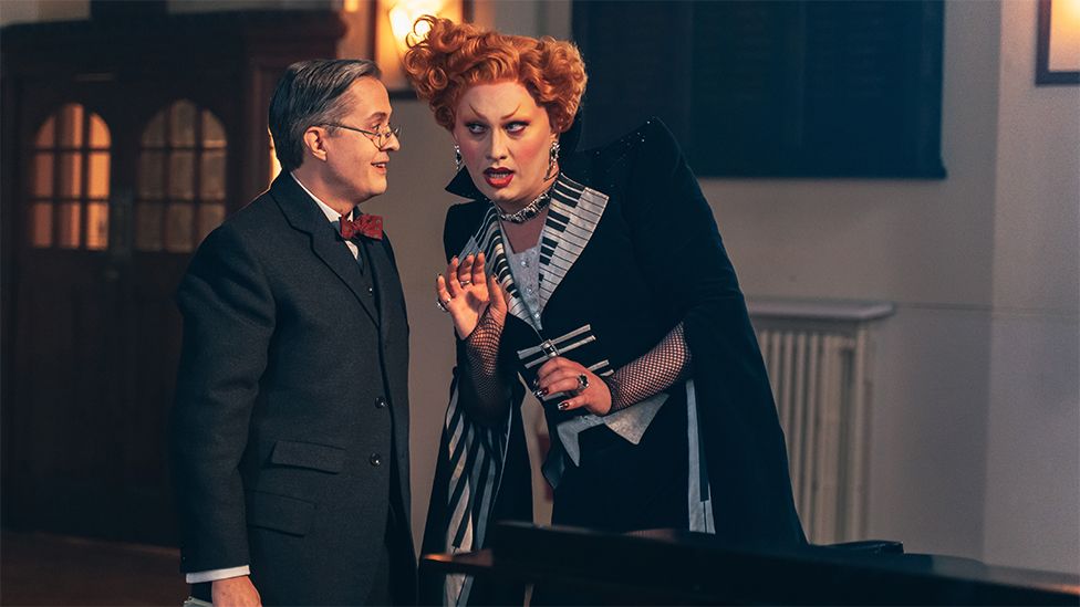 A man in an immaculately cut navy coloured suit complete with red bow tie speaks to a villainous character who wears a cape-style garment decorated with a piano key pattern on the oversized collar and interior lining. They have red hair in an Elizabethan perm, and wear various pieces of silver jewellery. They are leaning in towards the man, listening with intent. The scene has a slightly comical, pantomime feel.