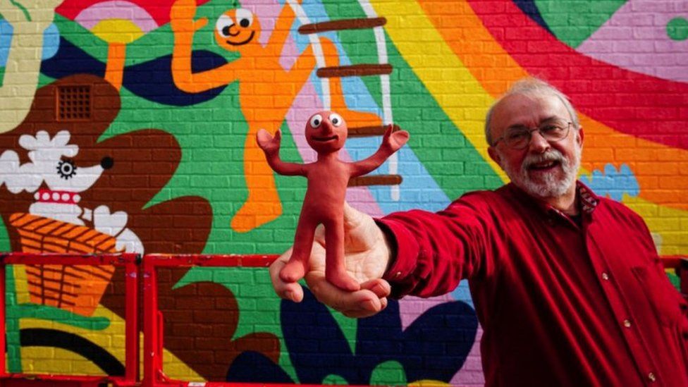 Peter Lord, the co-creator of Morph holding a Morph figurine. Peter is wearing glasses and a red jacket, stood in front of the mural. He is smiling and holding his arm stretched out towards the camera. The orange Morph figurine is in the centre of the frame. The figurine is positioned with both arms up above its head.