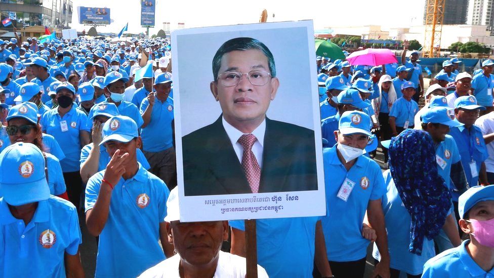 Толпа сторонников правящей партии в синей форме и кепках марширует в Пномпене 7 января в поддержку Хун Сена, один из них несет большой плакат с фотографией премьер-министра
