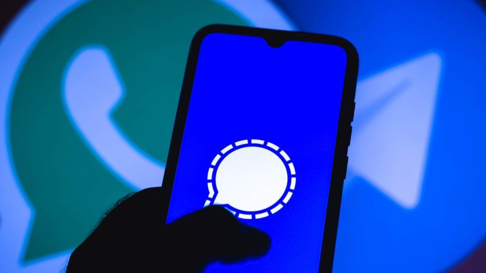 A person holds a phone with Signal's logo on it, in front of a WhatsApp logo