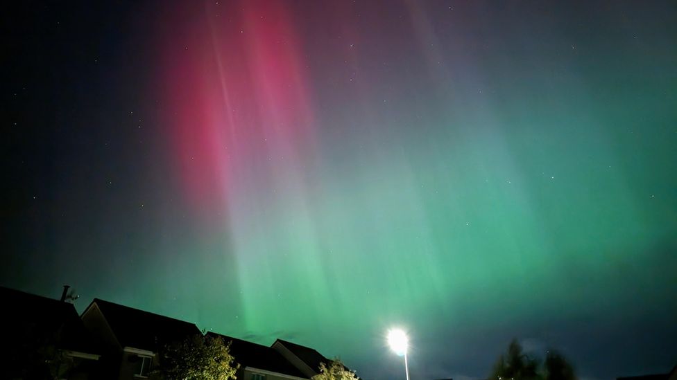 Northern Lights in turquoise and pink above the rooves of houses and a street light