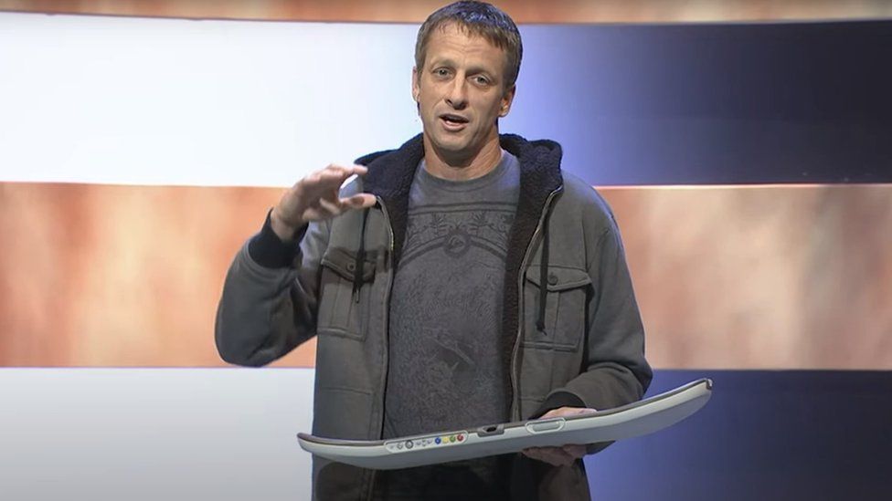 Skateboarder Tony Hawk wears a grey hoodie/jacket over the top of a grey/blue t-shirt. He's on a stage, and holding a plastic skateboard deck with no wheels. Along the side facing the audience there's a series of coloured buttons, including the x,y, a and b buttons from the standard Xbox controller