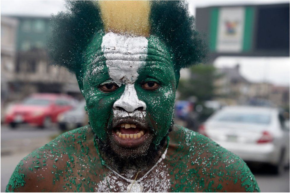 A man with his face painted in the colours of the Nigerian flag – green, white and green – staring into the camera. He is shirtless and outside.