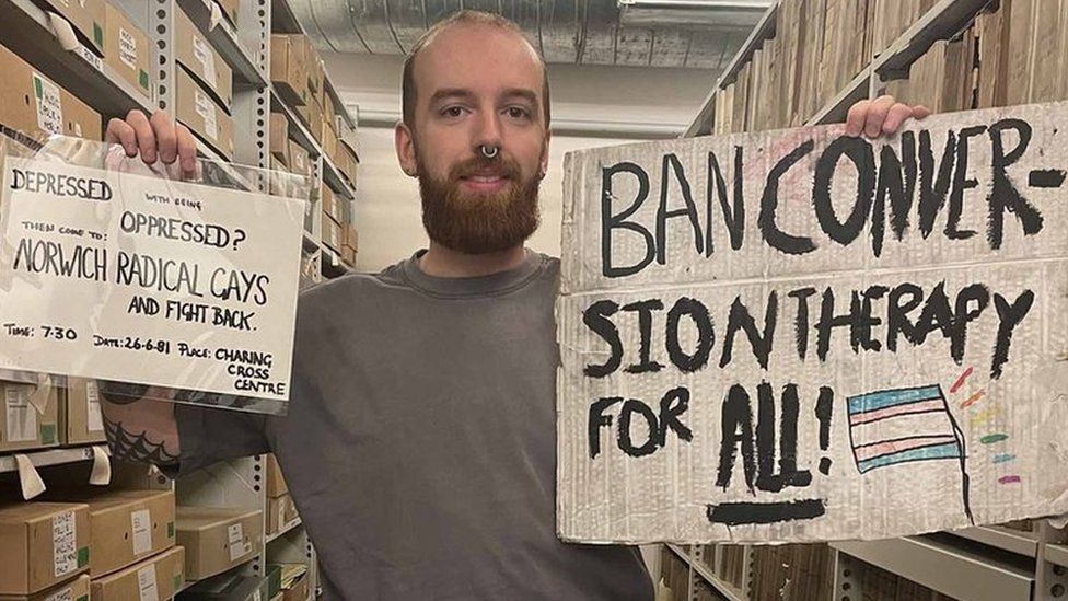 Adam Baker at Norfolk Heritage Centre holding up a poster and a sign that are in the Queer Norfolk archive