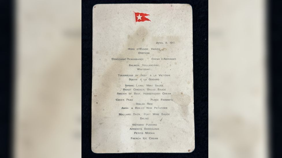 First-Class menu from the Titanic. It has water stains on it and a red emblem of a flag at the top.