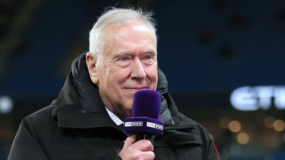 Football commentator Martin Tyler wearing a black coat and holding a blue microphone that says Bein Sports on it