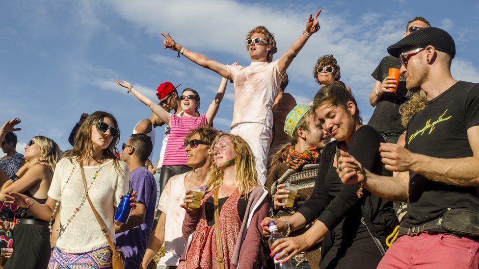 Несколько человек в яркой одежде поднимают руки , подпевая песням на фестивале Secret Garden Festival. Люди держат в руках напитки, бутылки с водой, носят солнцезащитные очки и шляпы, а солнце светит, а небо голубое с оттенком облаков.