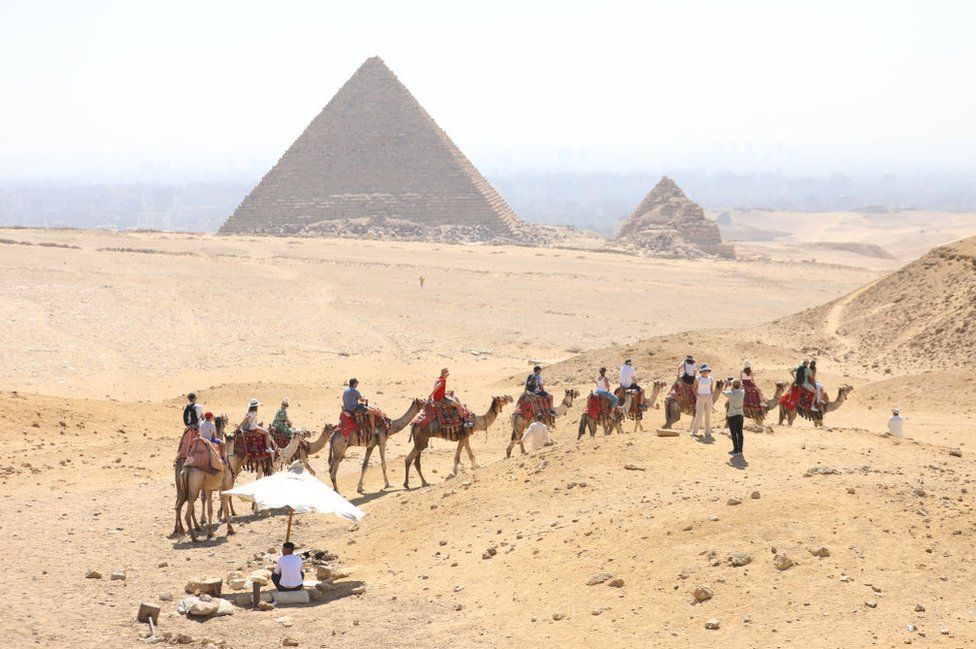 Two pyramids in the Egyptian surrounded by acres of sandy desert are visible in the background behind a haze of heat. The one on the left is very large, while the structure to its right is much smaller. In the foreground, a line of camels carrying tourists towards the attraction snakes across the frame. One couple appears to have dismounted, with a woman in a sunhat and white vest posing for a photo taken by her partner. In the bottom left corner of the frame a man shelters from the bright sun under a white parasol on a post driven into the sand.