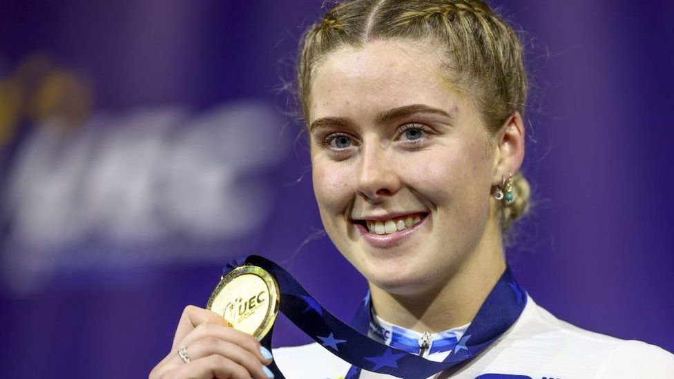 Winner Emma Finucane (GBR) with the gold medal during the ceremony of the final sprint on the third day of the European Track Cycling Championships in the Apeldoorn Omnisportcentrum. Emma is a 21-year-old white woman with blonde hair which she wears braided. She is dressed in a white sports jacket with blue stripes. She holds up a gold medal which she wears around her neck