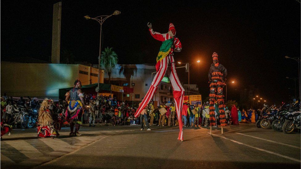 Two stilt walkers wearing colourful clothing with a crowd behind them