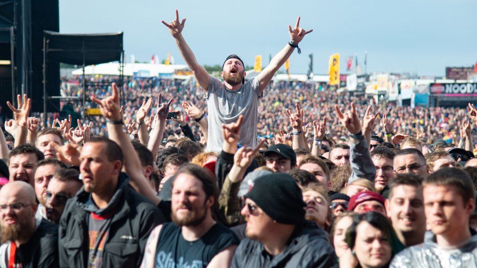 Festival goers cheering in the crowd during day 2 of Download Festival 2019. A white man with a beard sits above the crowd on another spectator's shoulders, his hands stretched up making the sign of horns. The hand gesture, associated with rock music, is repeated by other members of the crowd. The audience is outside on a sunny day and in the background flags and food outlets can be seen.