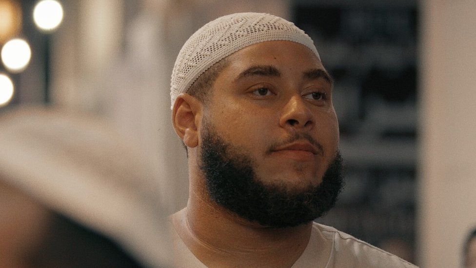 Big Zuu from his his documentary Big Zuu Goes to Mecca. Big Zuu is a 28-year-old black man with shaved hair, a dark beard and brown eyes. He's pictured in traditional ihram clothing - white robes and a white skull cap.
